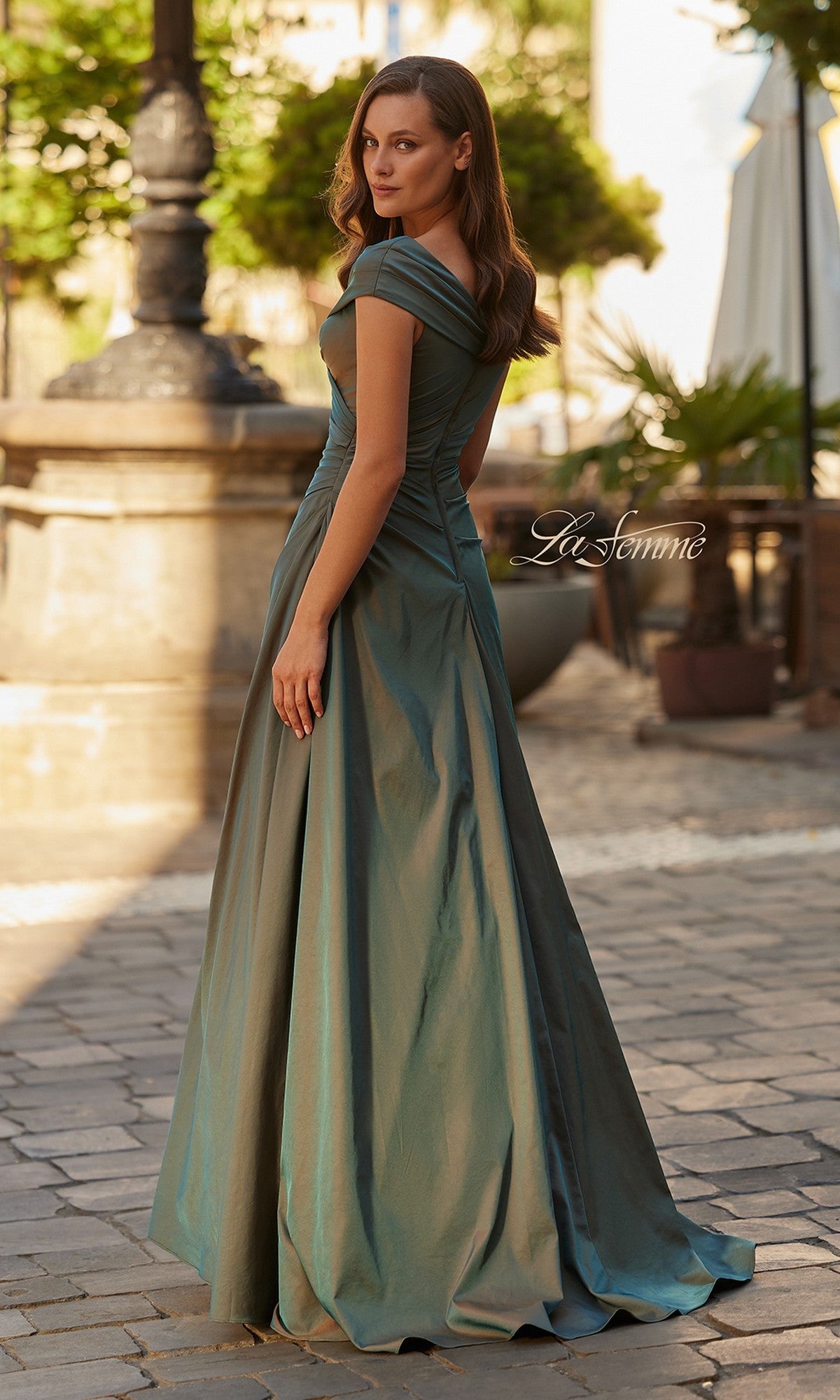 A woman in the La Femme 33446 long formal dress stands on a cobblestone street, surrounded by charming outdoor seating and lush greenery.